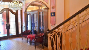 A staircase with ornate metal railings, a chandelier, and a red table with chairs in a room with tiled flooring and a large window. - Renesance Krasna Kralovna (Karlovy Vary)