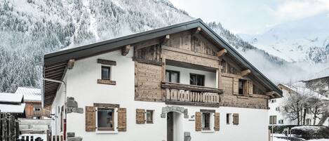 Un chalet de dos pisos con balcones de madera, base de piedra y montañas al fondo.