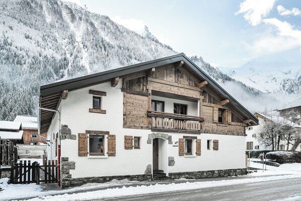 A two-story chalet with wooden balconies, a stone foundation, and a mountain backdrop.