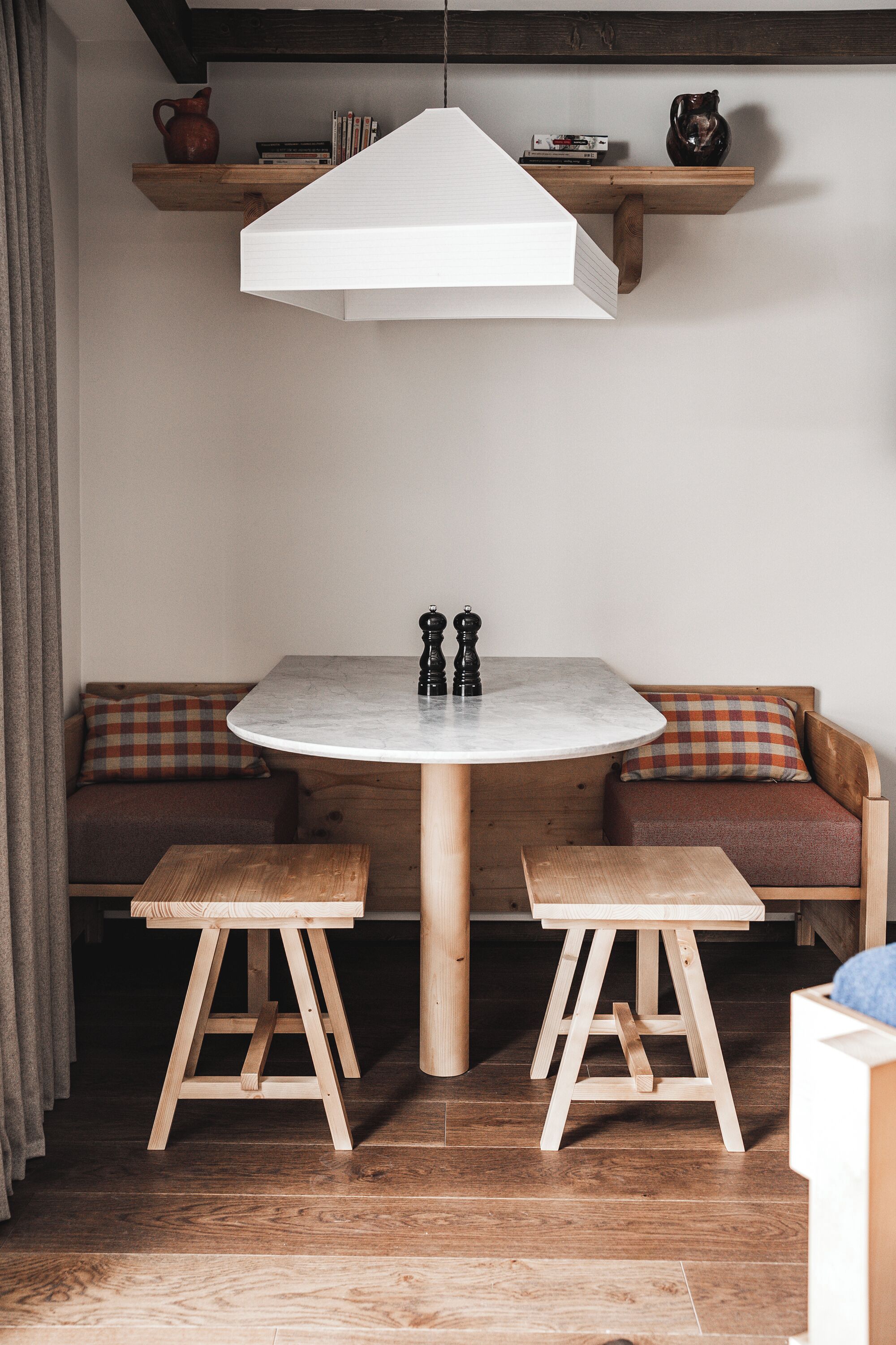 Comedor con una mesa redonda, dos bancos de madera, una lámpara colgante blanca y un estante de madera arriba.