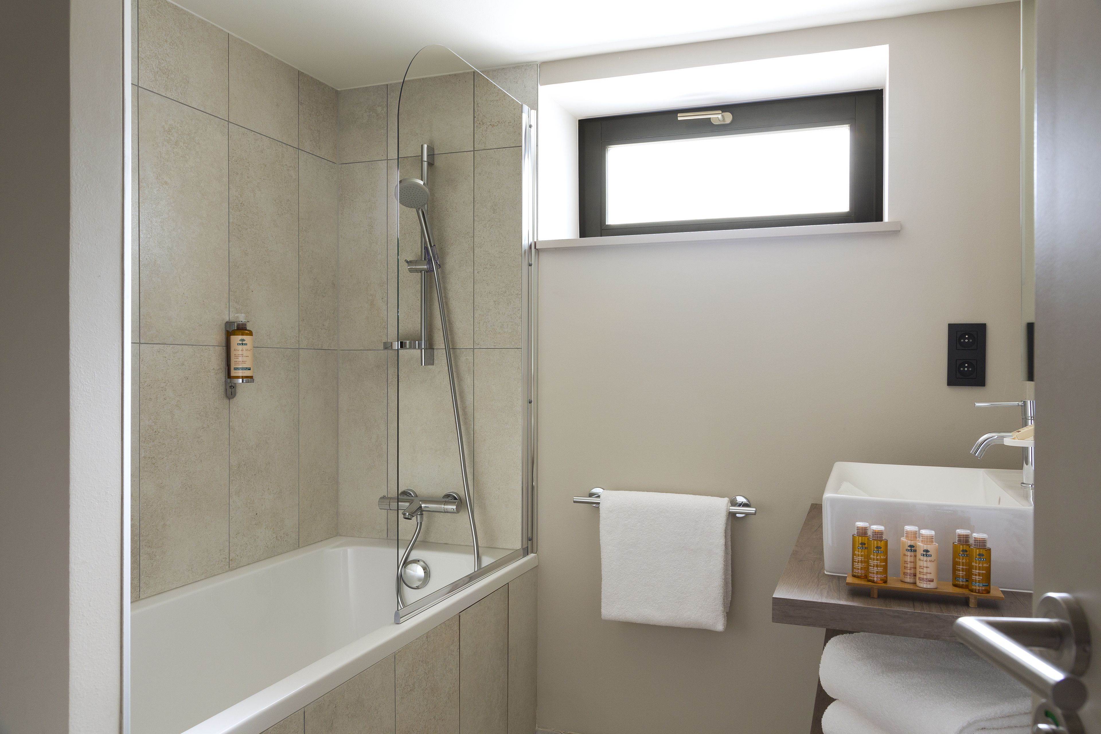 Ein modernes Badezimmer mit Badewanne, einer Glaskabine für die Dusche, einem Fenster über dem Waschbecken und Pflegeprodukten auf einem Regal.
