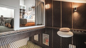 Ein modernes Hotelzimmer mit einem großen Fenster, einem Bett mit kariertem Kopfkissen, einem kleinen Hocker und einer Wandleuchte.