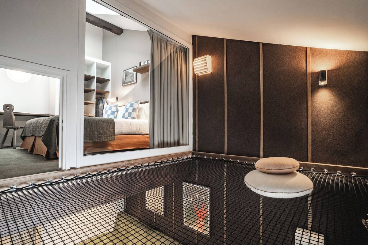 Et moderne hotelværelse med et stort vindue, en seng med pude i tern, en lille skammel og en vægmonteret lampe.