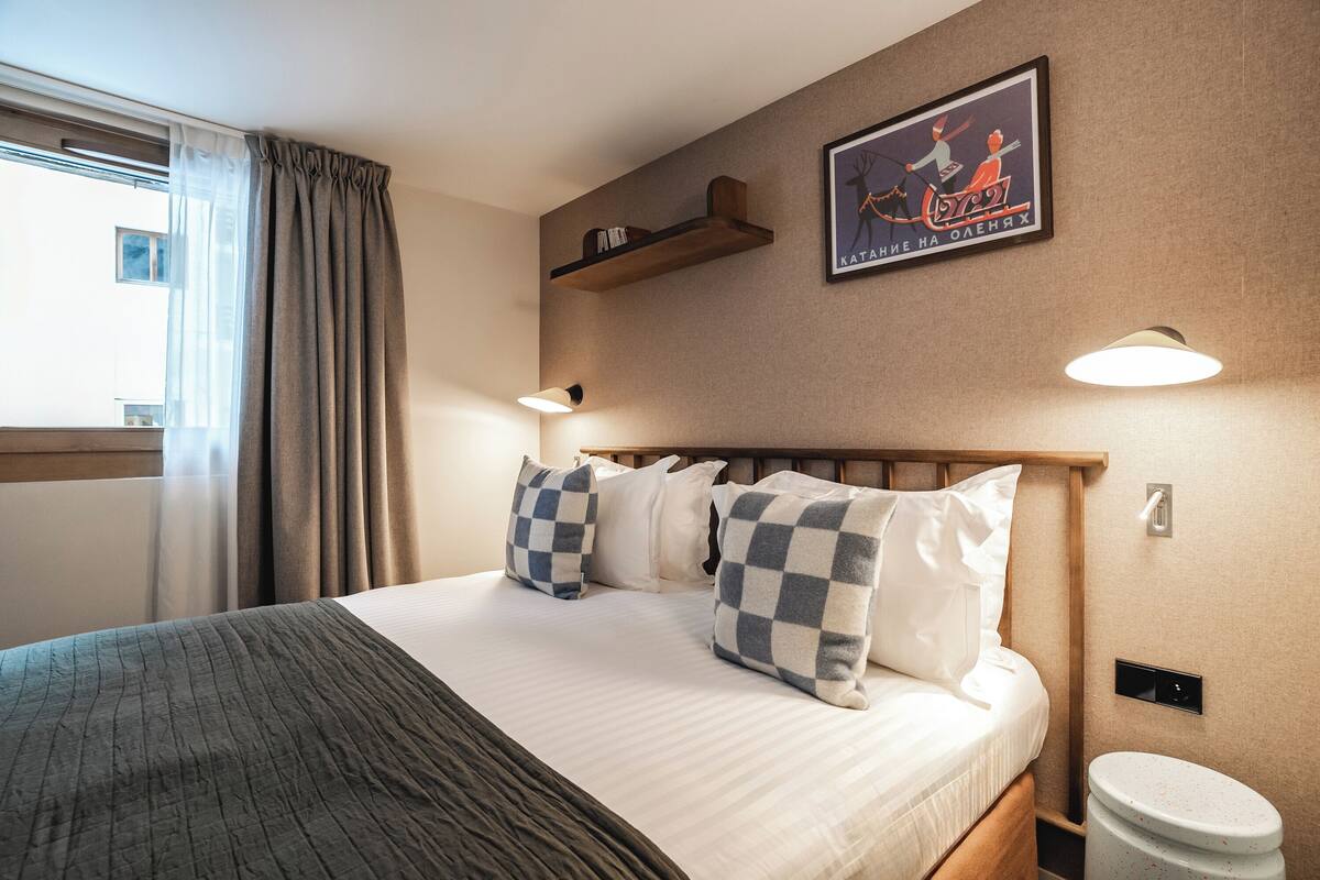 En pænt redt seng med ternede puder, et sengebord med en lampe, en vægmonteret hylde med bøger og et indrammet plakat.