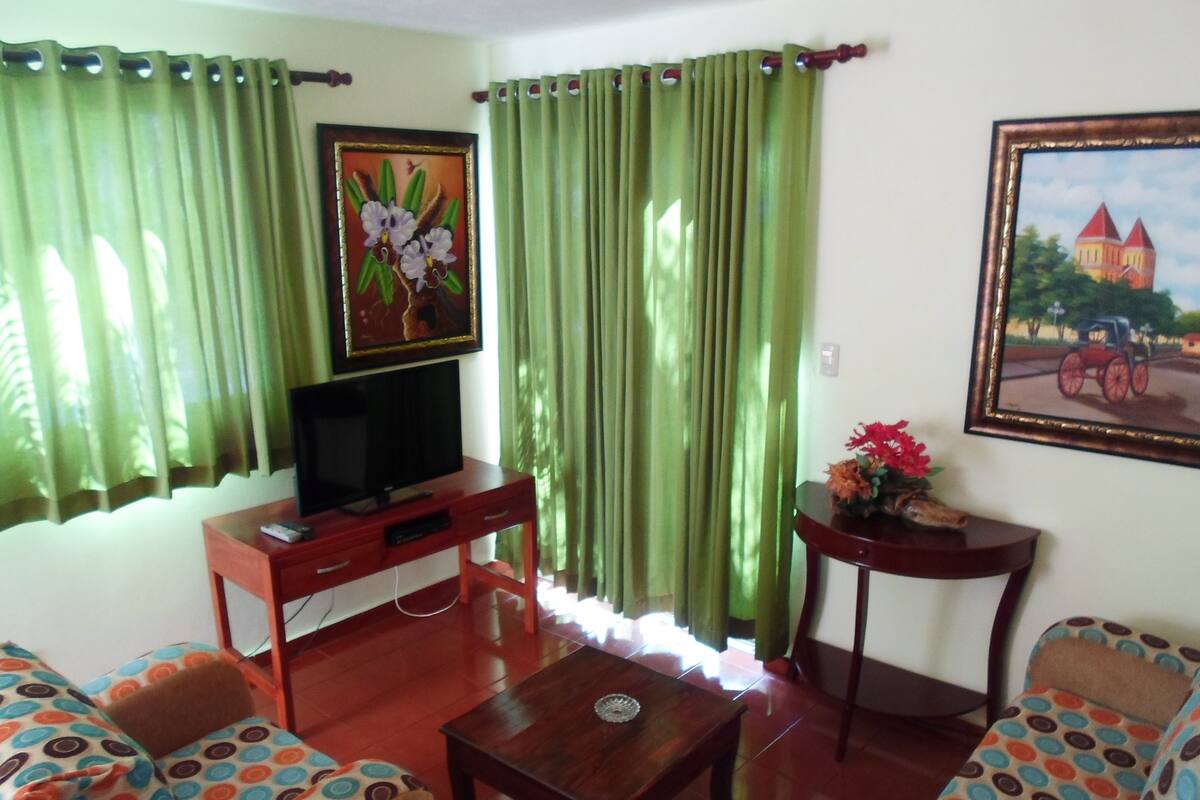 A living room with a TV on a wooden stand, green curtains, a painting, and a small table with a vase.