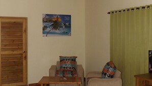 Una habitación con techo de madera, una cama, un sillón con cojines coloridos, una mesita y un cuadro en la pared.