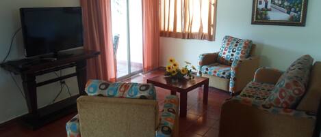 Una sala de estar con televisor, dos sofás estampados, una mesa de centro con flores y un cuadro en la pared.
