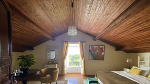 Une chambre à coucher avec un plafond en bois, un lit avec un pied de lit vert, une table de chevet, une chaise, un bureau avec un miroir, une plante en pot et une fenêtre avec des rideaux.