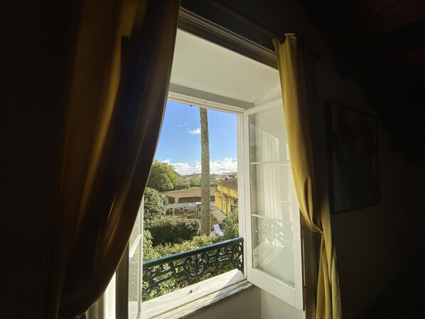 Una ventana con cortinas, un balcón con barandilla metálica y vista a un edificio y árboles.