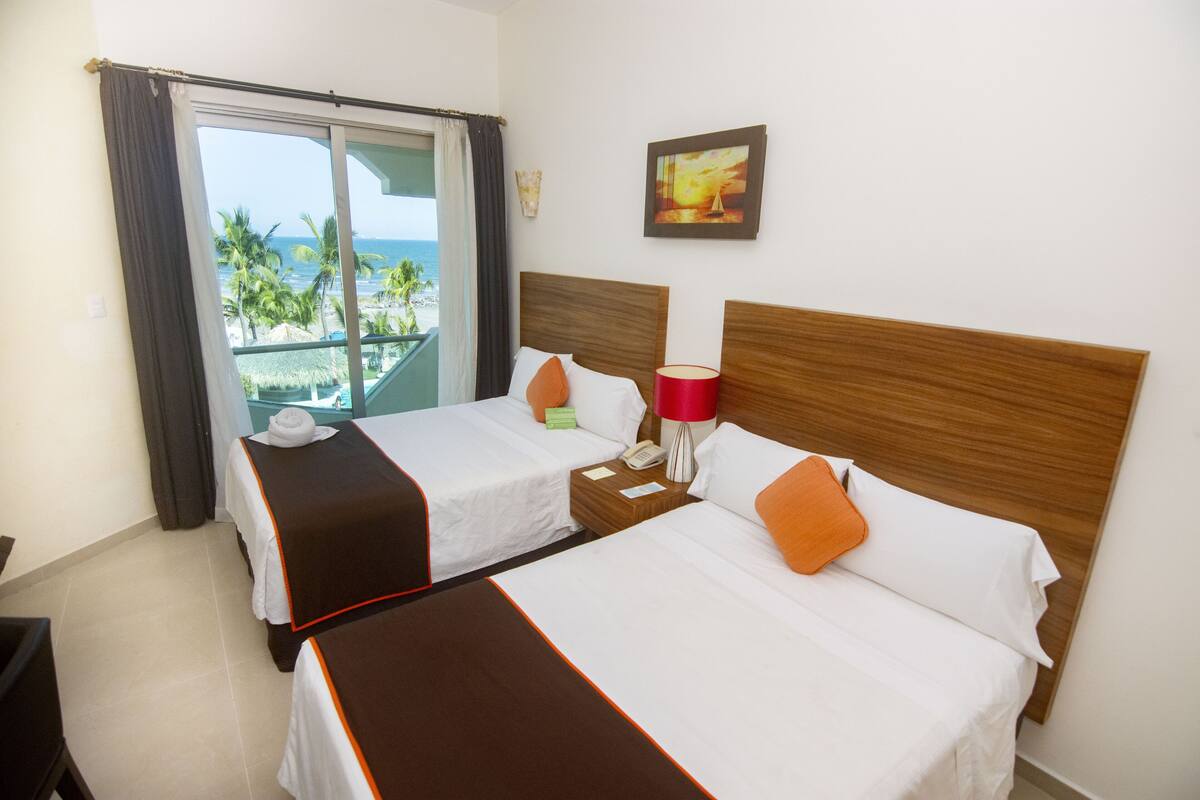 A hotel room with two beds, a balcony with a view of the ocean, and a painting on the wall.