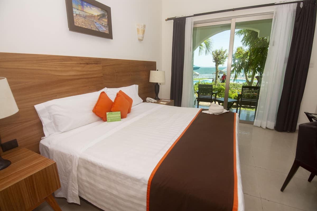 A hotel room with a bed, a nightstand, a lamp, a view of the ocean through a glass door, and a chair.