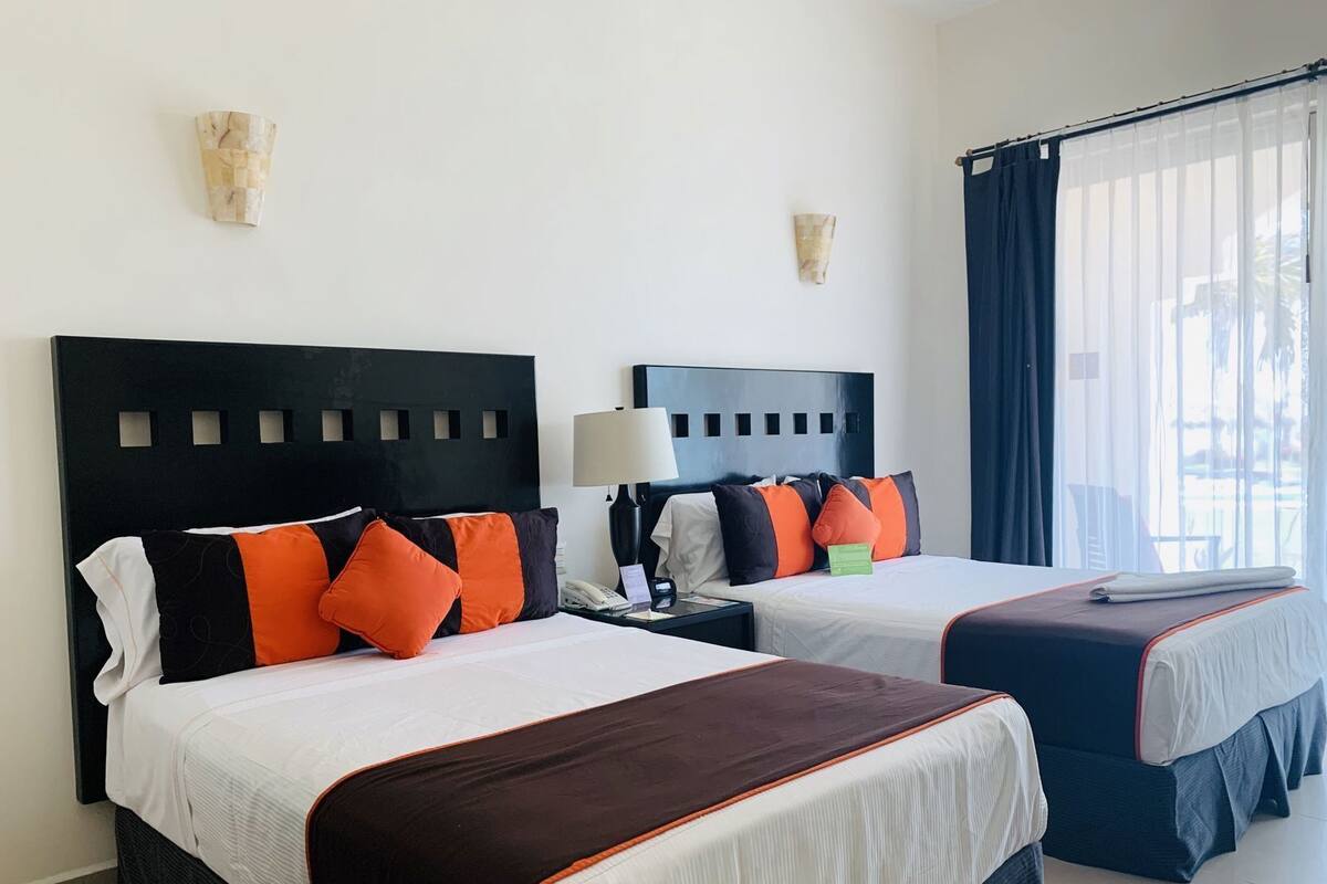 A hotel room with two beds, each with a black headboard, white bedding, and orange pillows. There is a nightstand with a lamp, a window with curtains, and a wall-mounted light fixture.