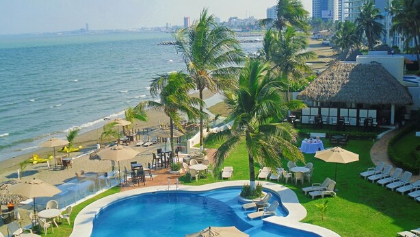 Una piscina de resort con sillas de descanso y sombrillas, un área de playa con sillas y sombrillas, y palmeras.
