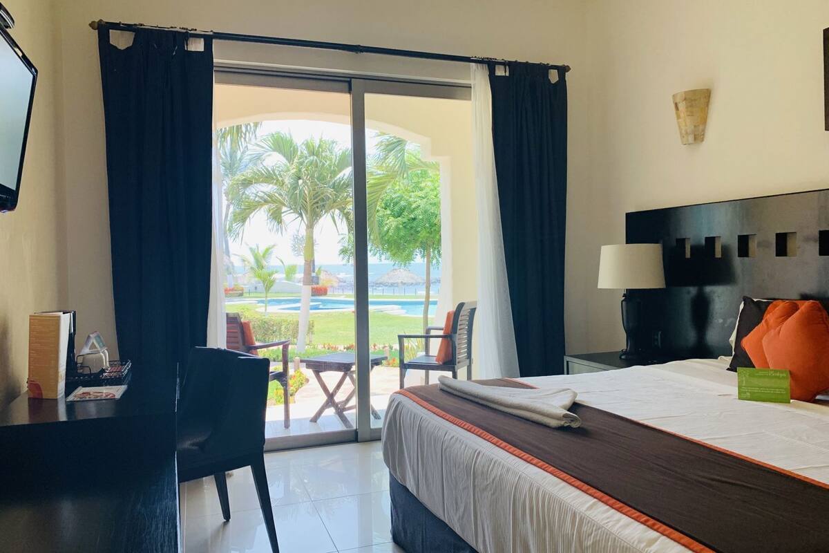 A hotel room with a bed, a desk, a chair, a TV, and a view of a pool and palm trees.