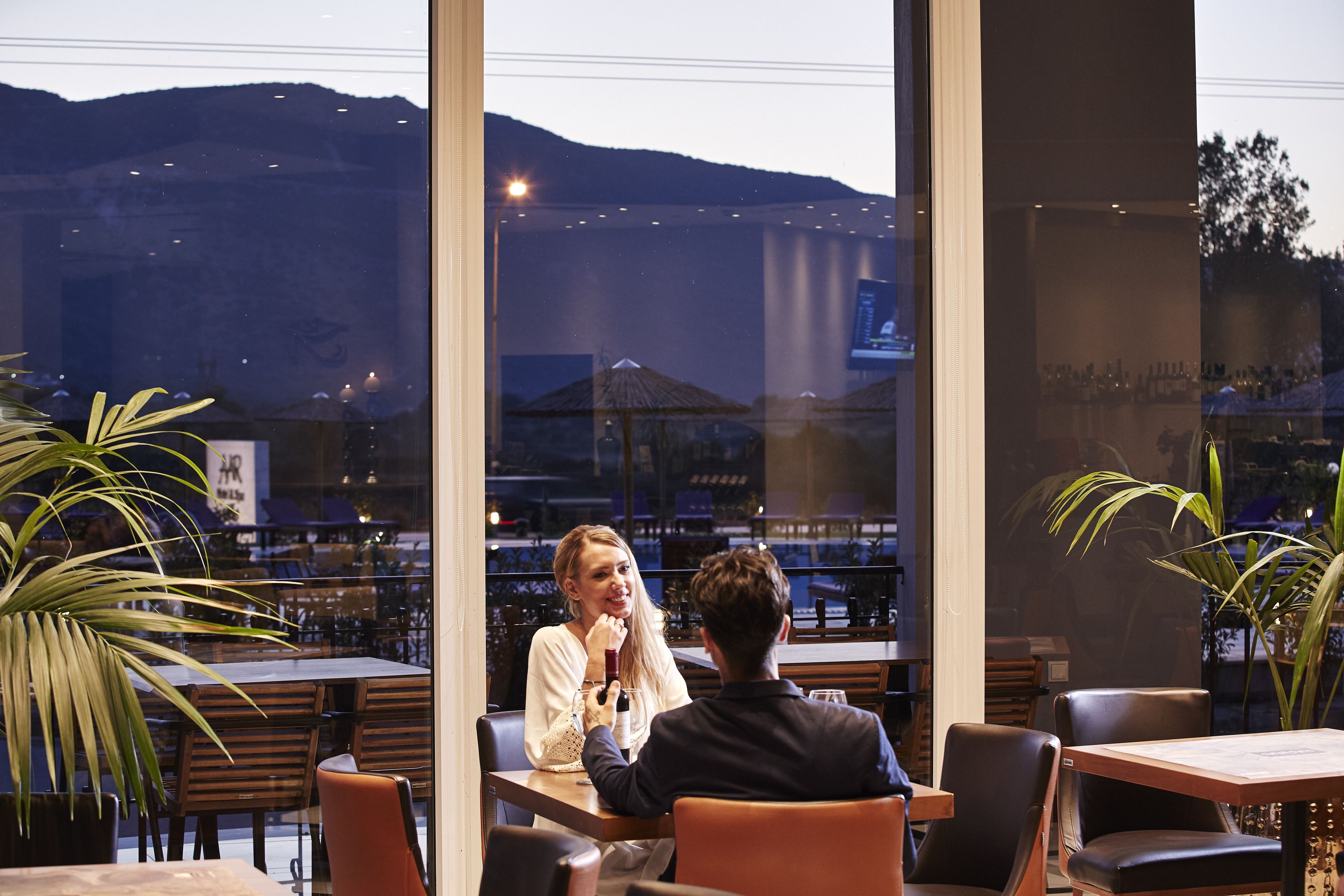 An indoor dining area with large windows, tables, chairs, and potted plants.