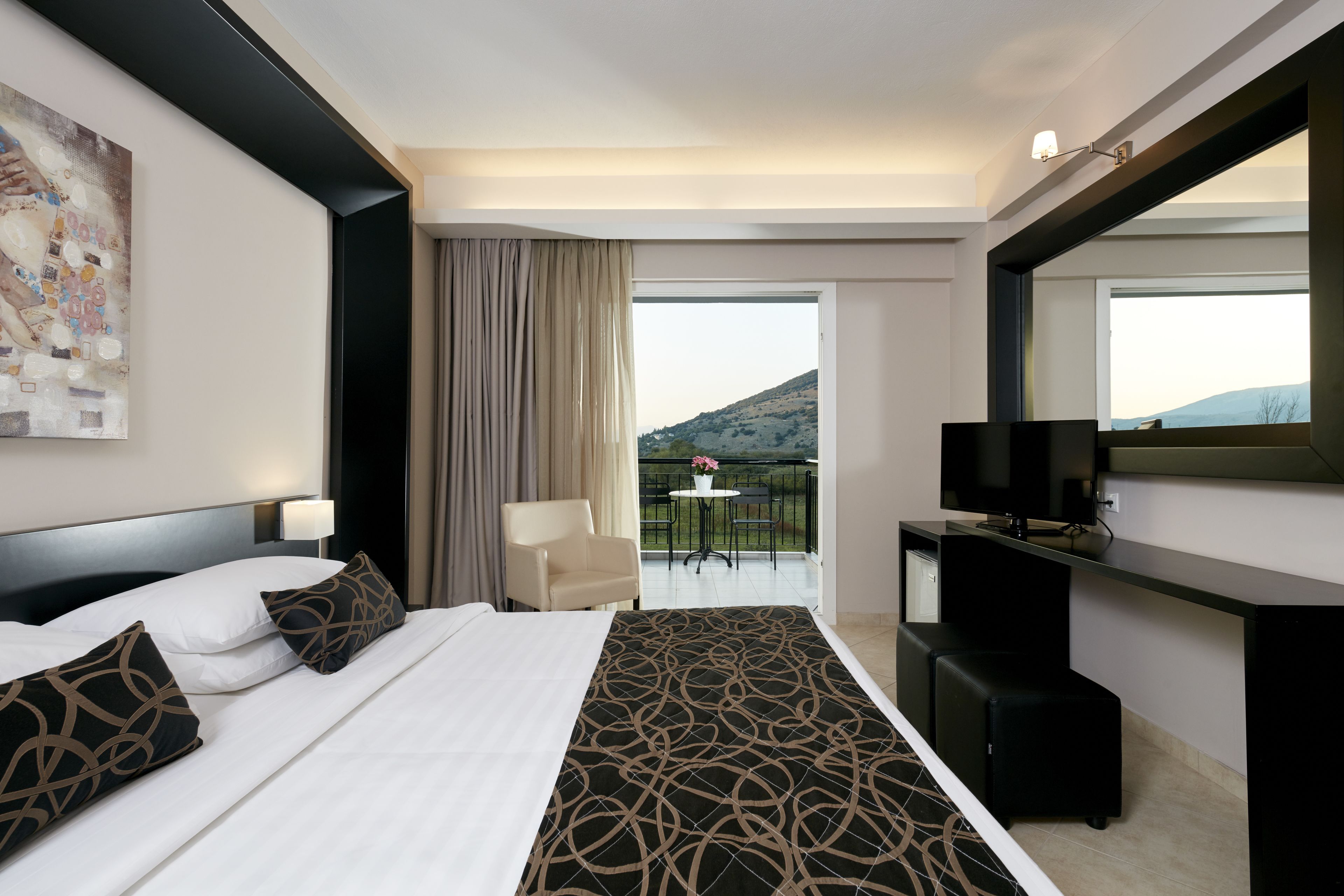 A hotel room with a bed, a desk, a television, a chair, and a balcony with a view of the hills.