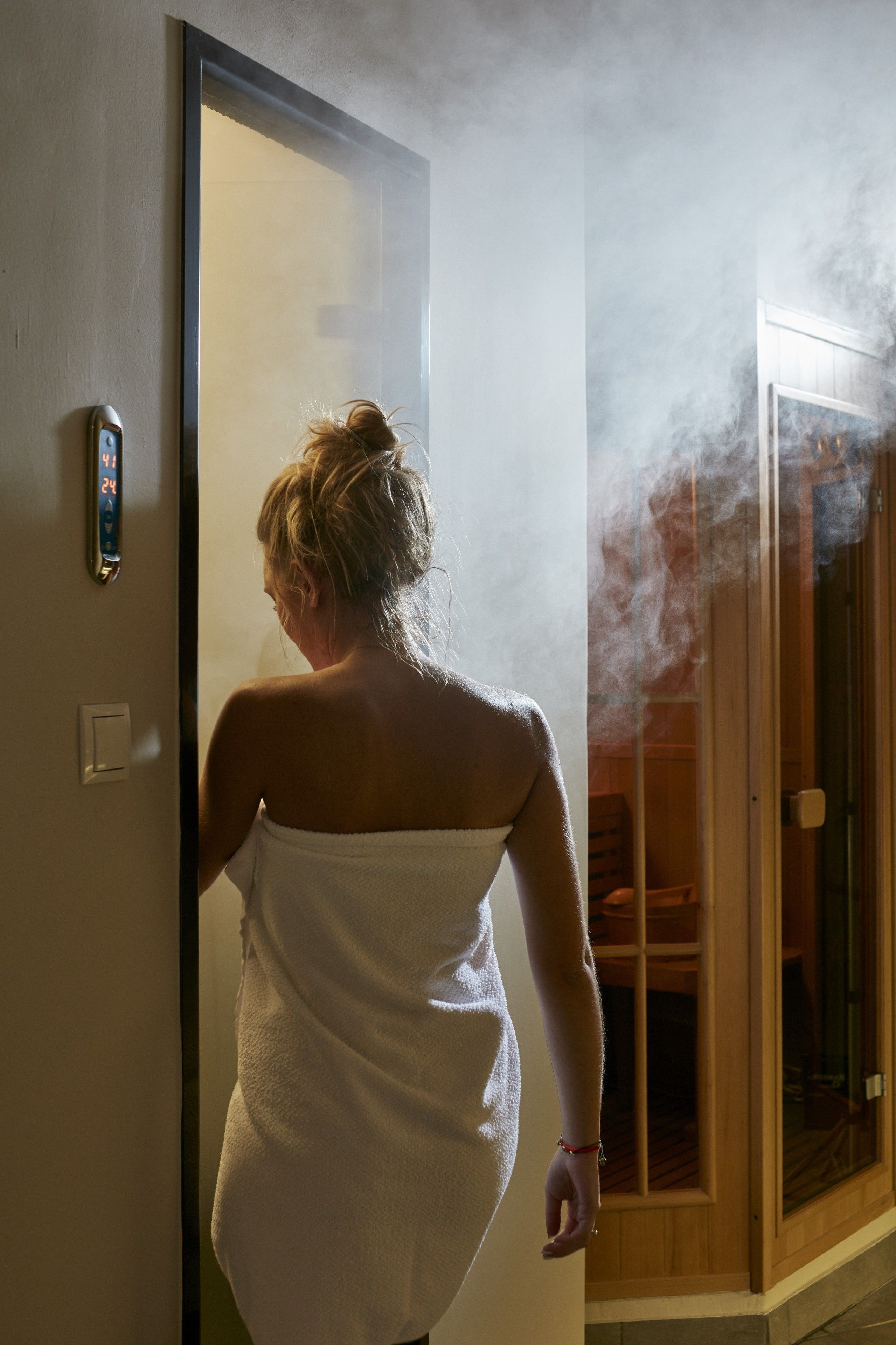 A person in a white towel standing at a doorway with steam visible, suggesting a sauna or steam room.