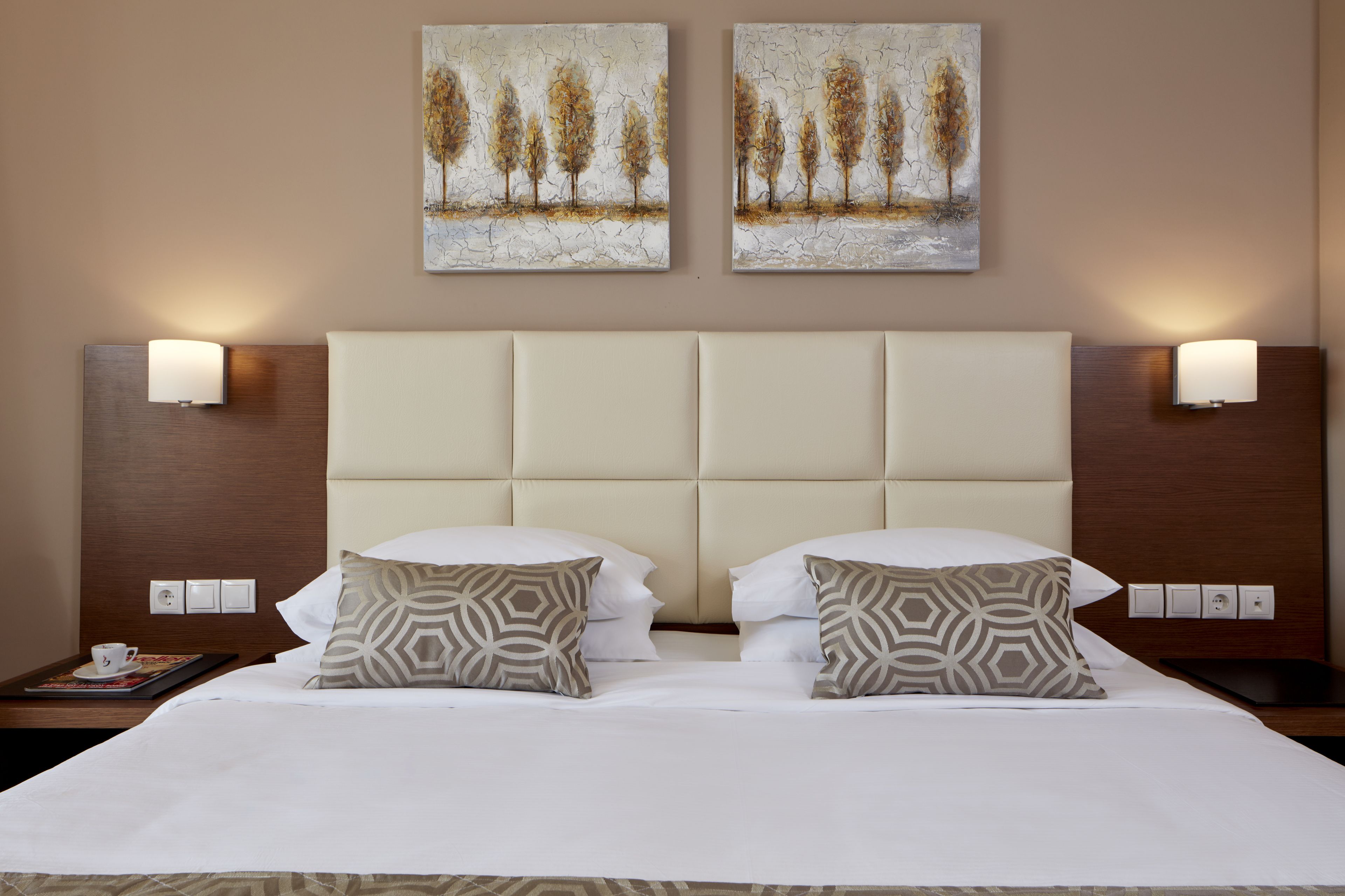A hotel room with a large bed, two wall-mounted lamps, and two paintings above the bed.