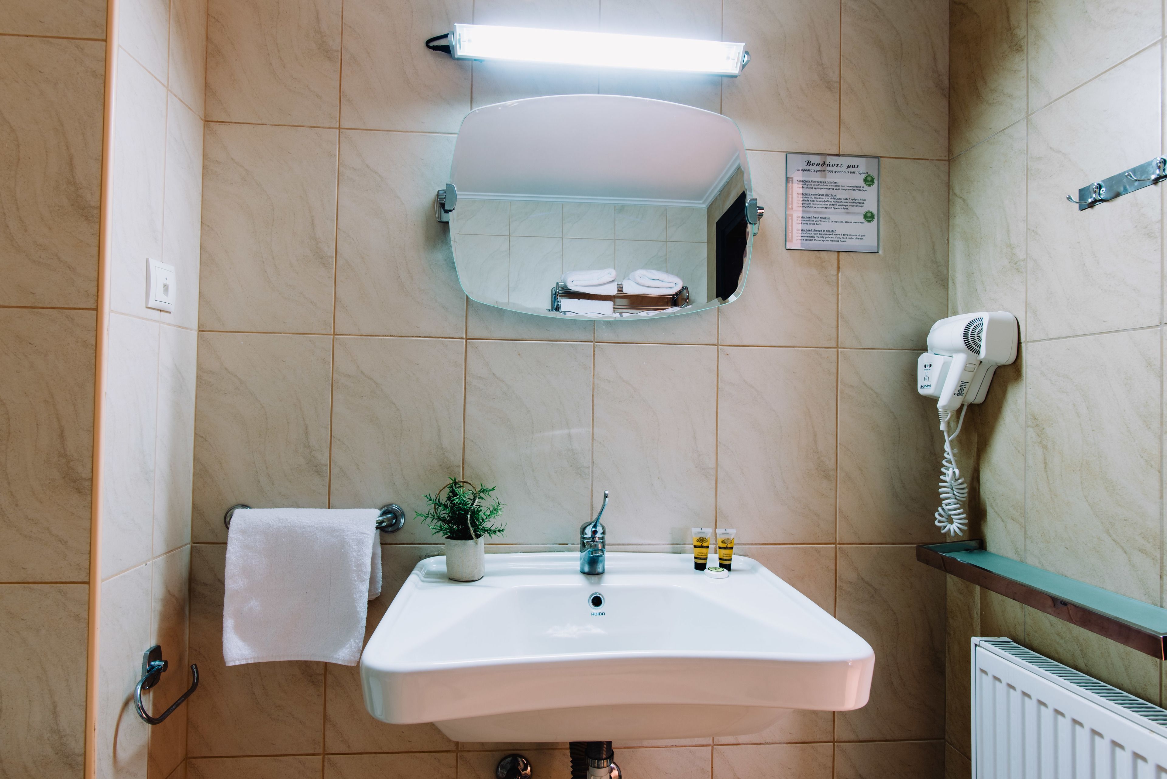 A bathroom with a white sink, a mirror, a towel rack, a small potted plant, a hairdryer, and a sign with instructions.