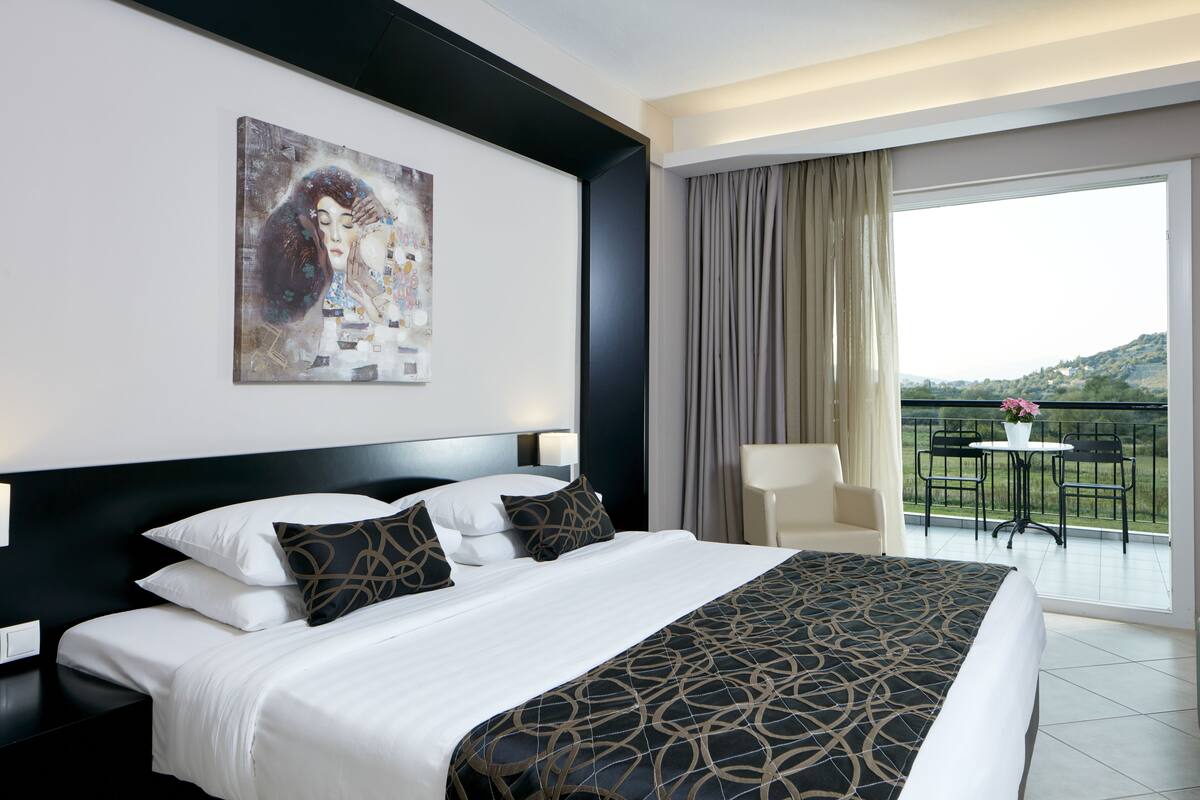 A hotel room with a large bed, a painting on the wall, a balcony with a table and chairs, and a view of the outdoors.