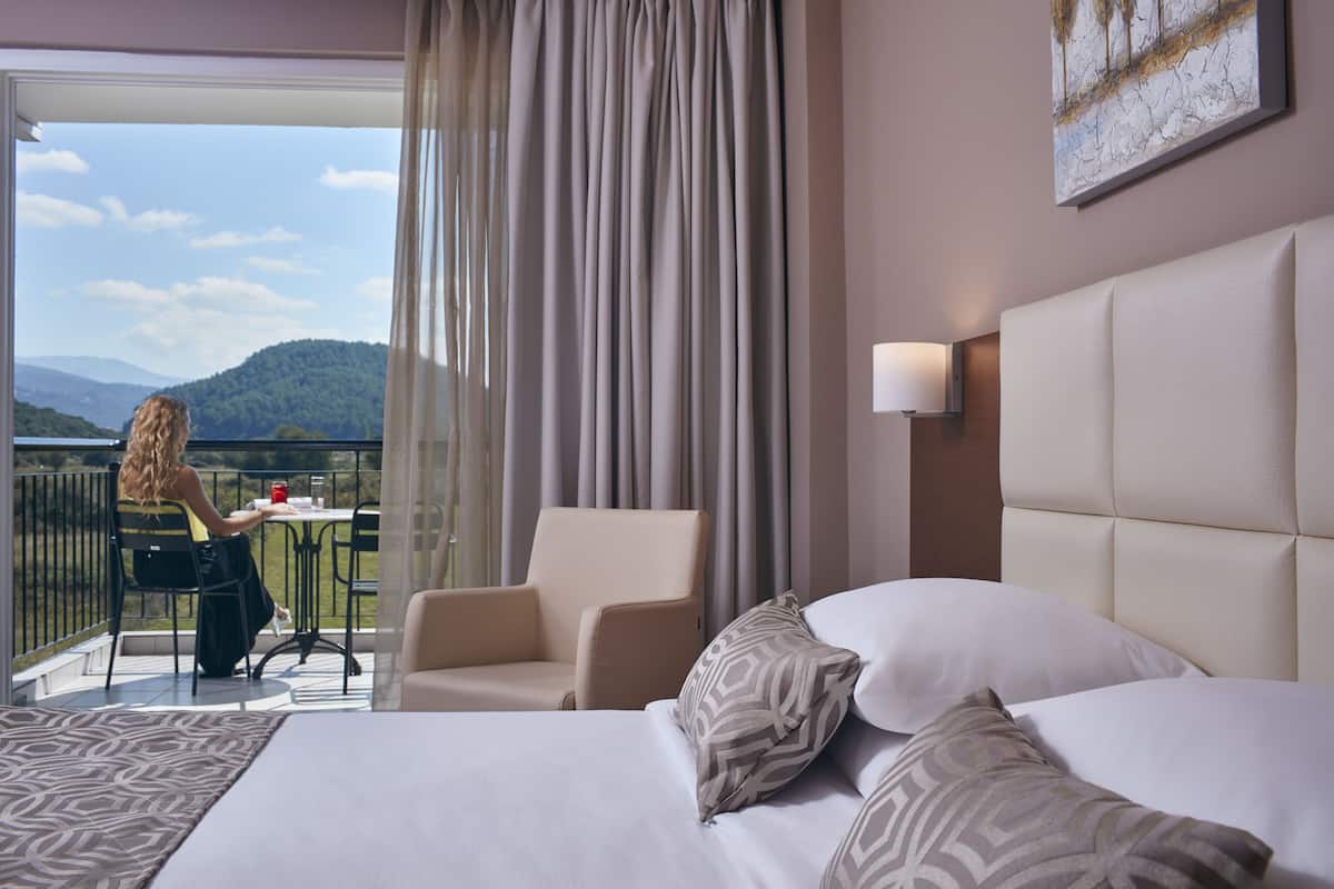 A hotel room with a bed, a chair, a table, and a balcony with a view of the mountains.