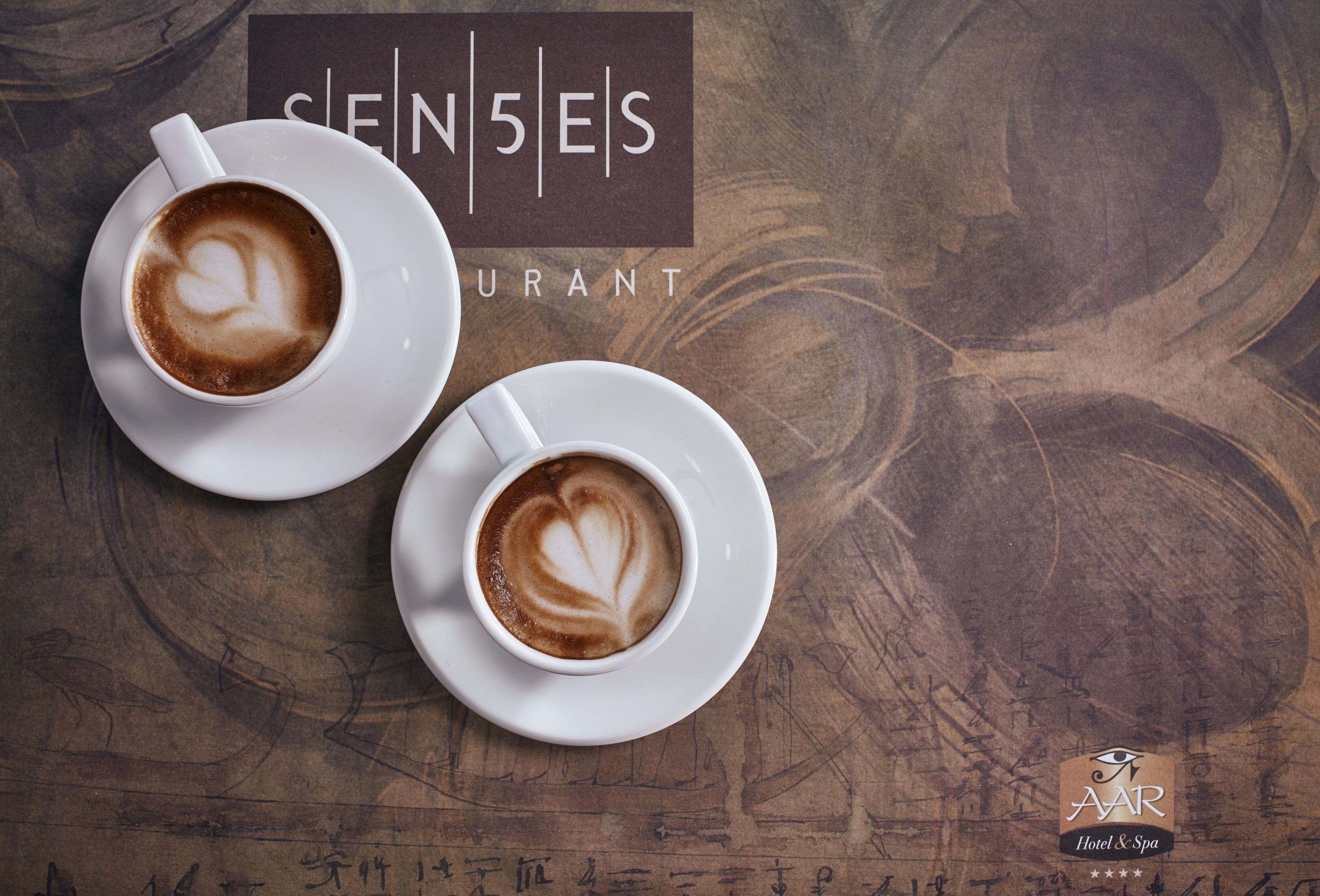 Two cups of coffee with latte art on white saucers, placed on a wooden table with a sign reading "SENSES RESTAURANT" in the background.