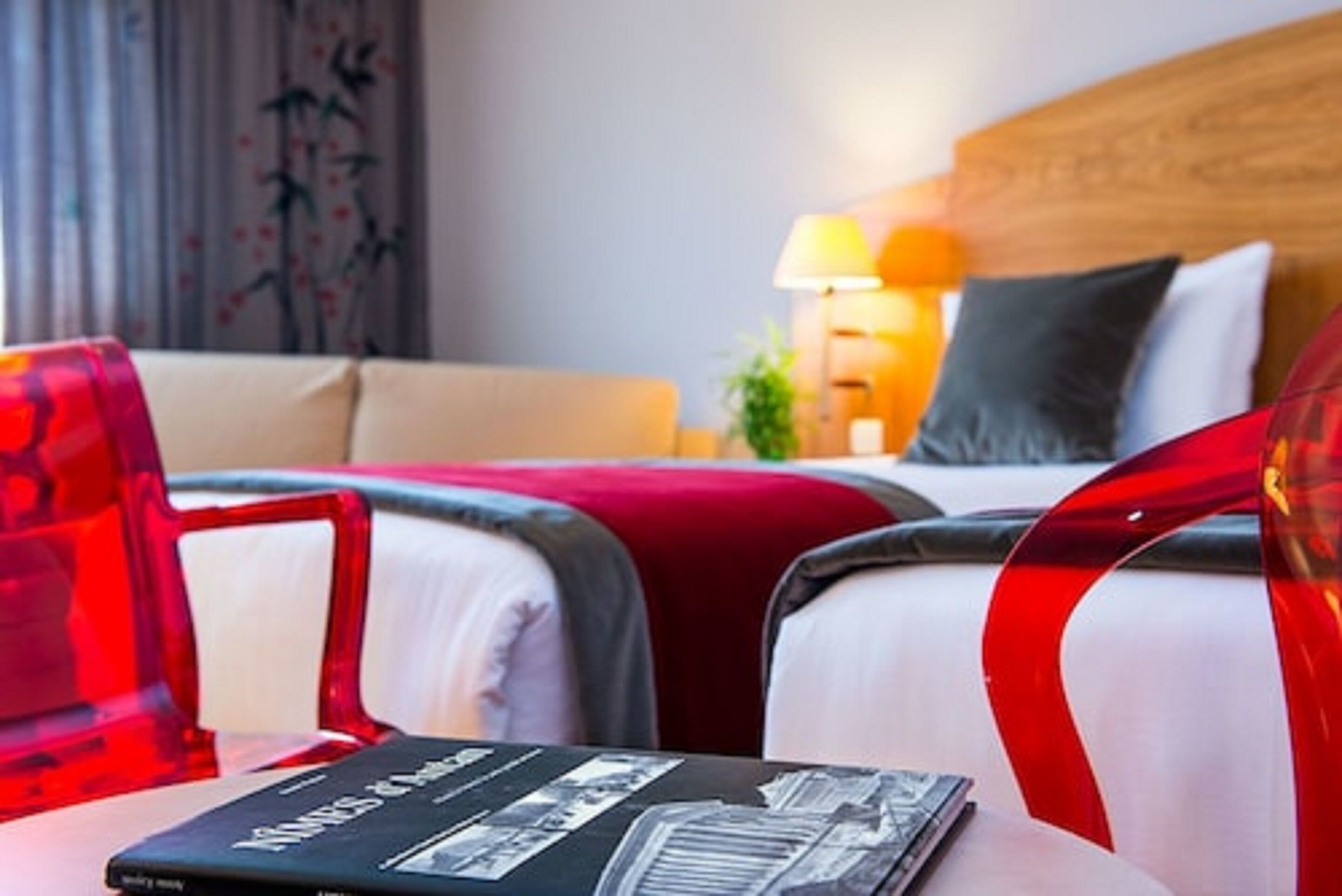 A hotel room with a bed, two red chairs, a table with a book, a lamp, and a plant.