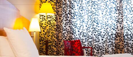 Una habitación de hotel con una cama, dos lámparas de noche, un sillón rojo y papel tapiz con motivos florales.