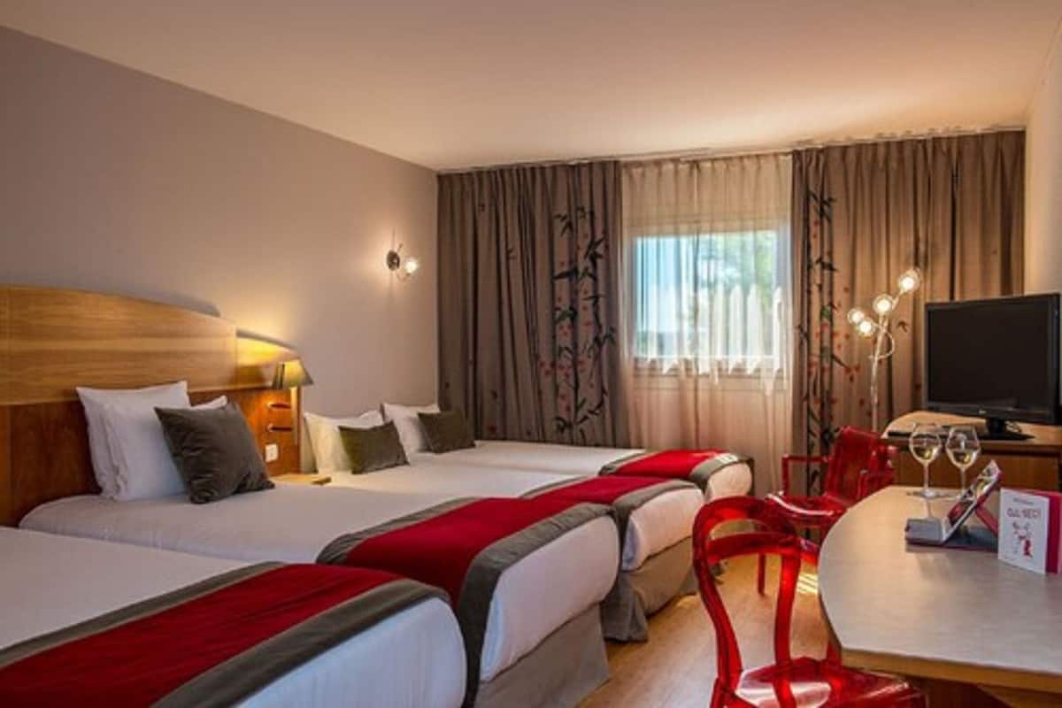 A hotel room with two beds, a TV, a dining table with chairs, and a window with curtains.