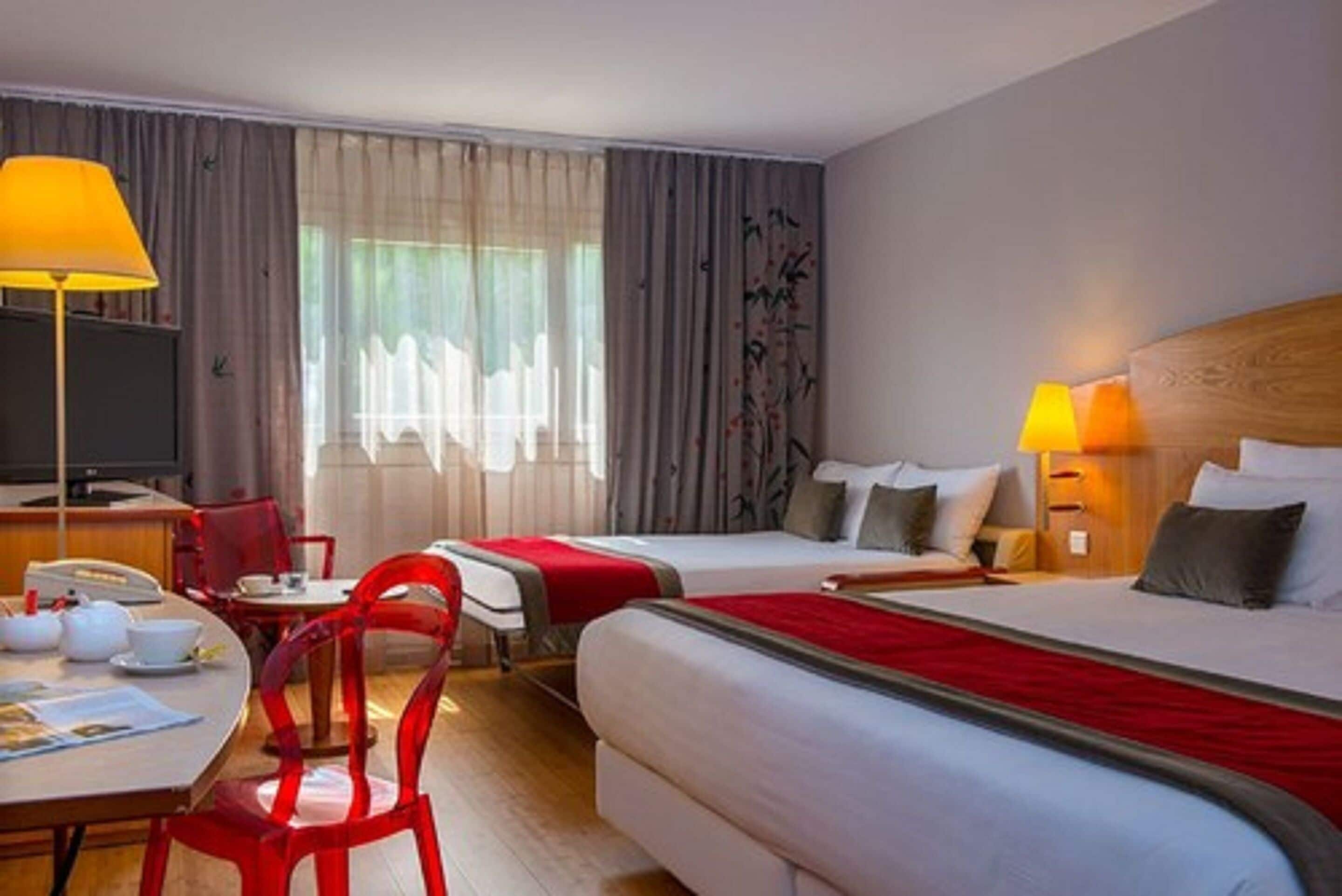 A hotel room with two beds, a desk with a chair, a TV, and a window with curtains.