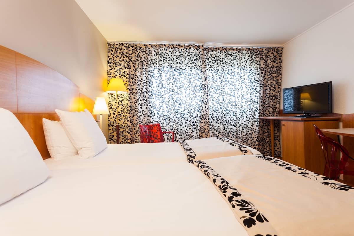 A hotel room with a bed, a desk, a television, and a window with floral curtains.