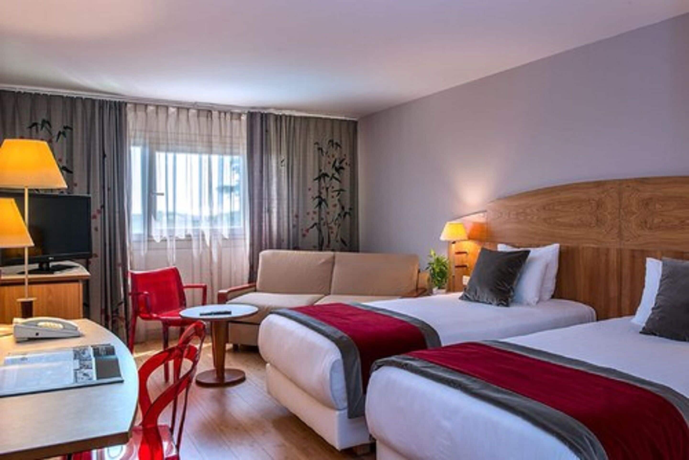 A hotel room with two beds, a sofa, a TV, and a desk with a lamp.