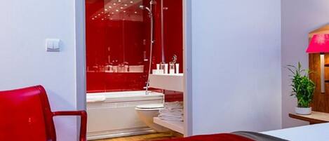 A modern hotel room with a red accent wall, a large bed, a red chair, a small table with a plant, and a bathroom with a red shower area.