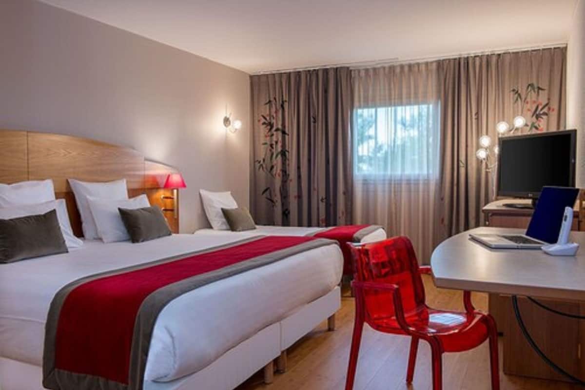 A hotel room with two beds, a desk with a laptop, a red chair, and a TV.
