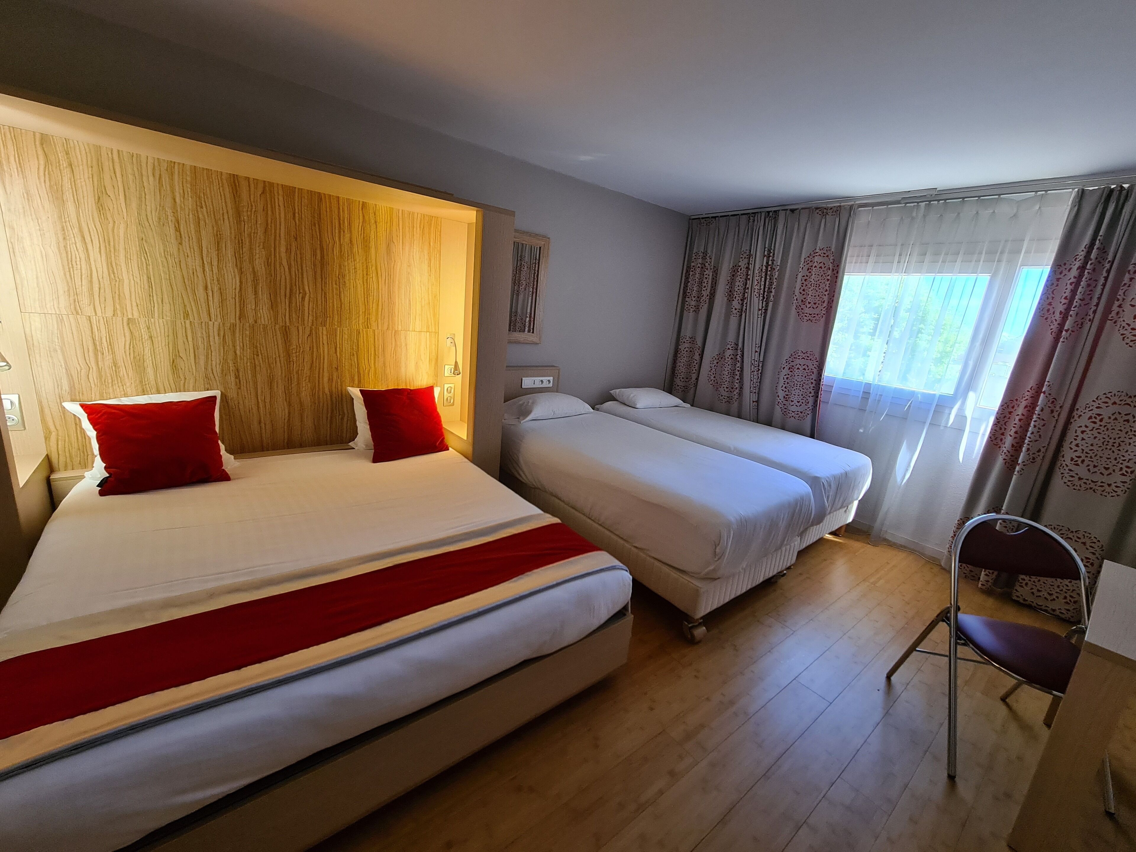 A hotel room with two beds, a wooden headboard, a chair, and a window with curtains.