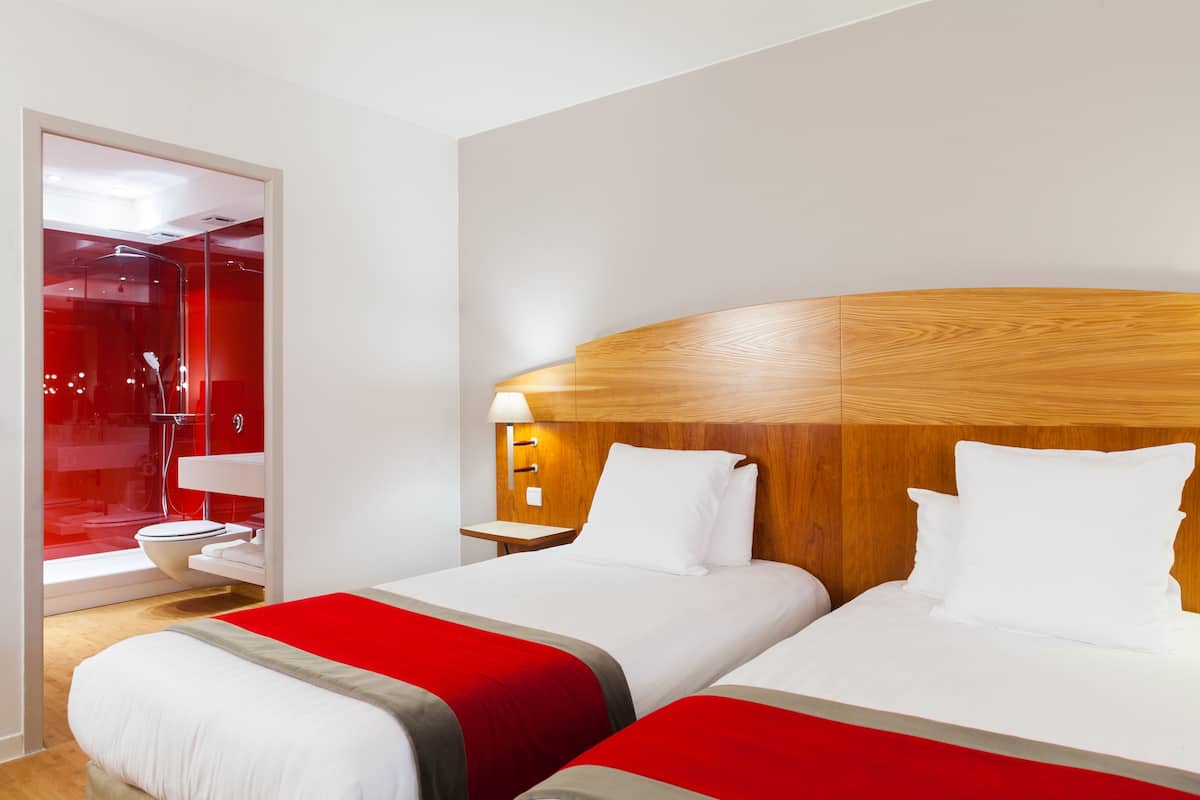 A hotel room with two beds, a wooden headboard, a red accent wall, and a glass shower enclosure.