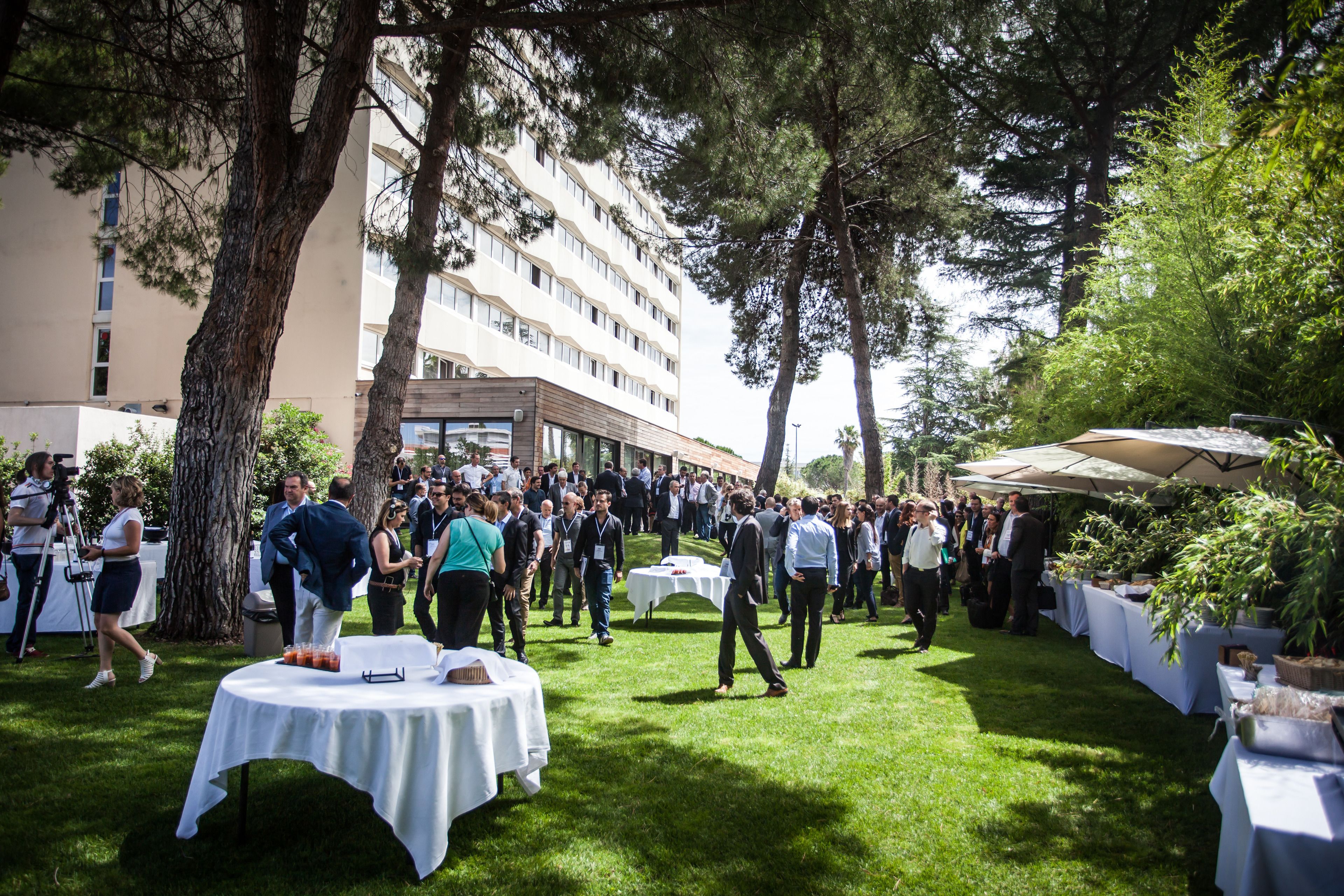 An outdoor event with people gathered around tables and chairs, a building in the background, and trees providing shade.