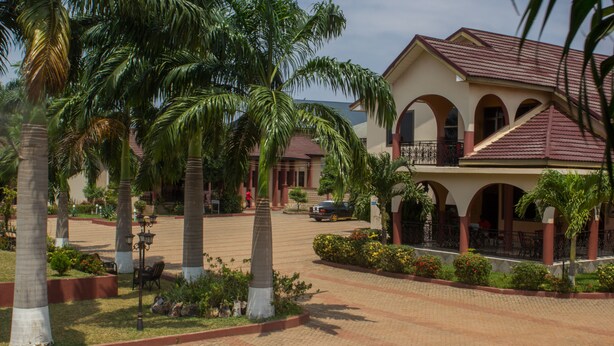 A building with a red-tiled roof, multiple arches, and a paved courtyard with palm trees and plants.