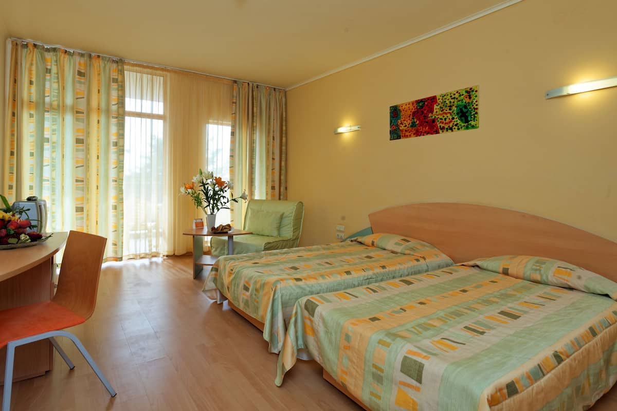 A hotel room with two beds, a desk, a chair, a table with a vase of flowers, and a painting on the wall.