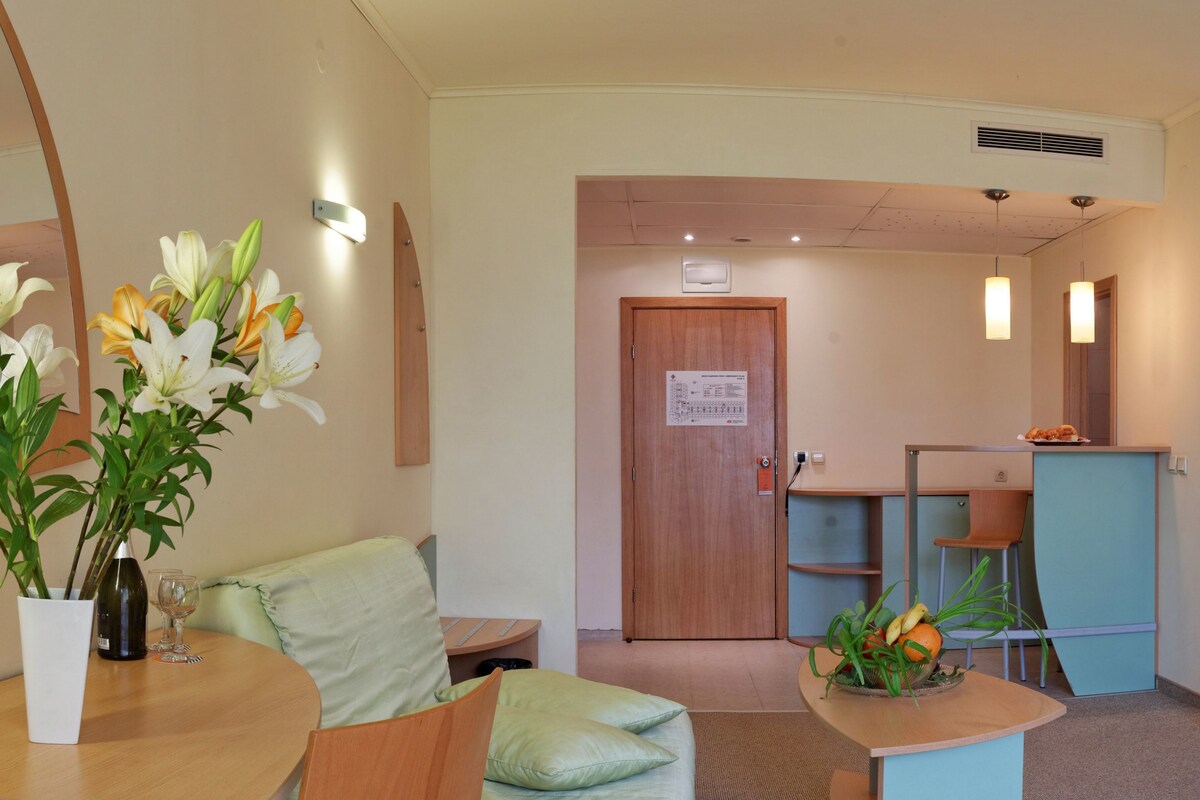 A hotel room with a wooden bed, a small desk with a chair, a white vase with flowers, and a wooden door with a sign.