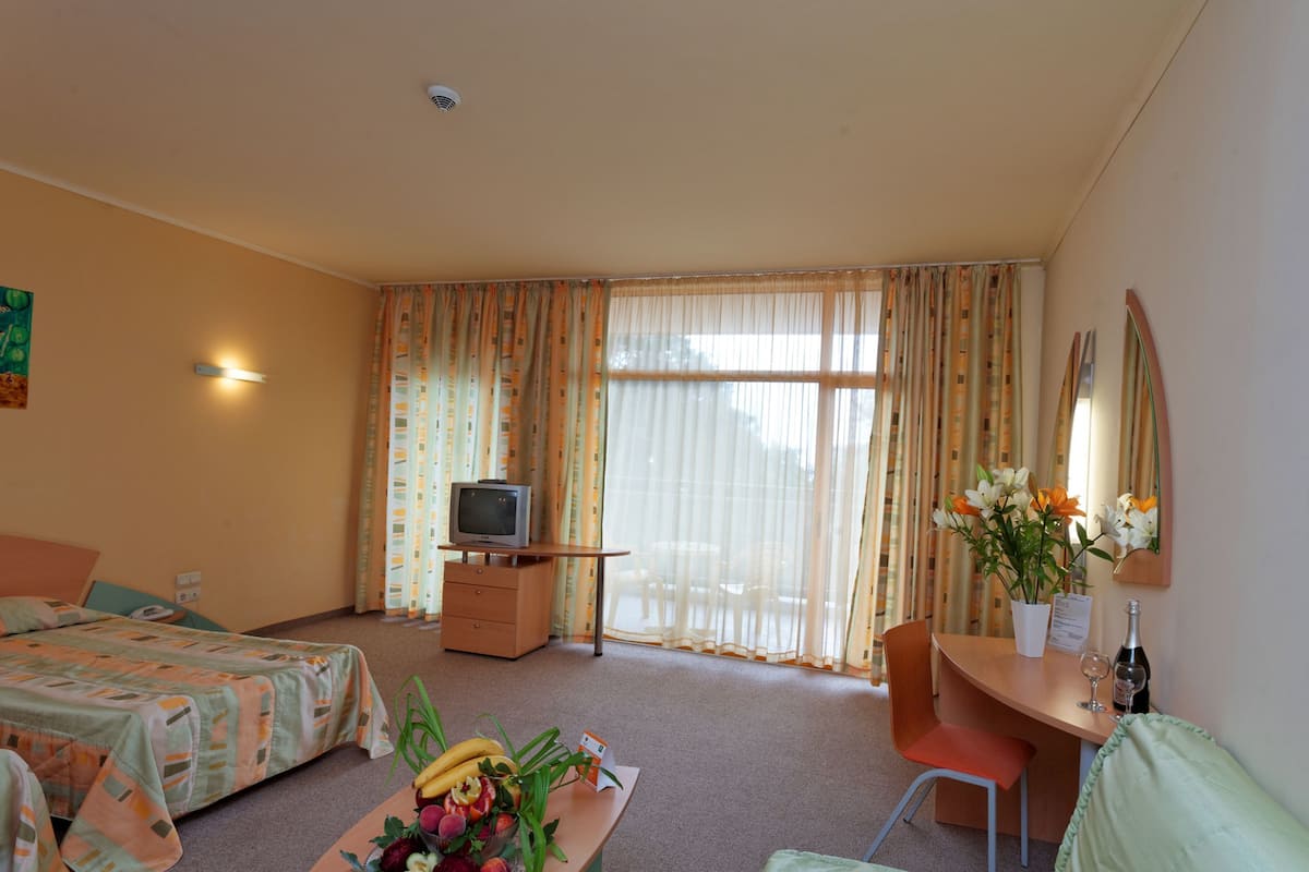 A hotel room with a bed, a desk with a chair, a television, and a table with fruits and flowers.