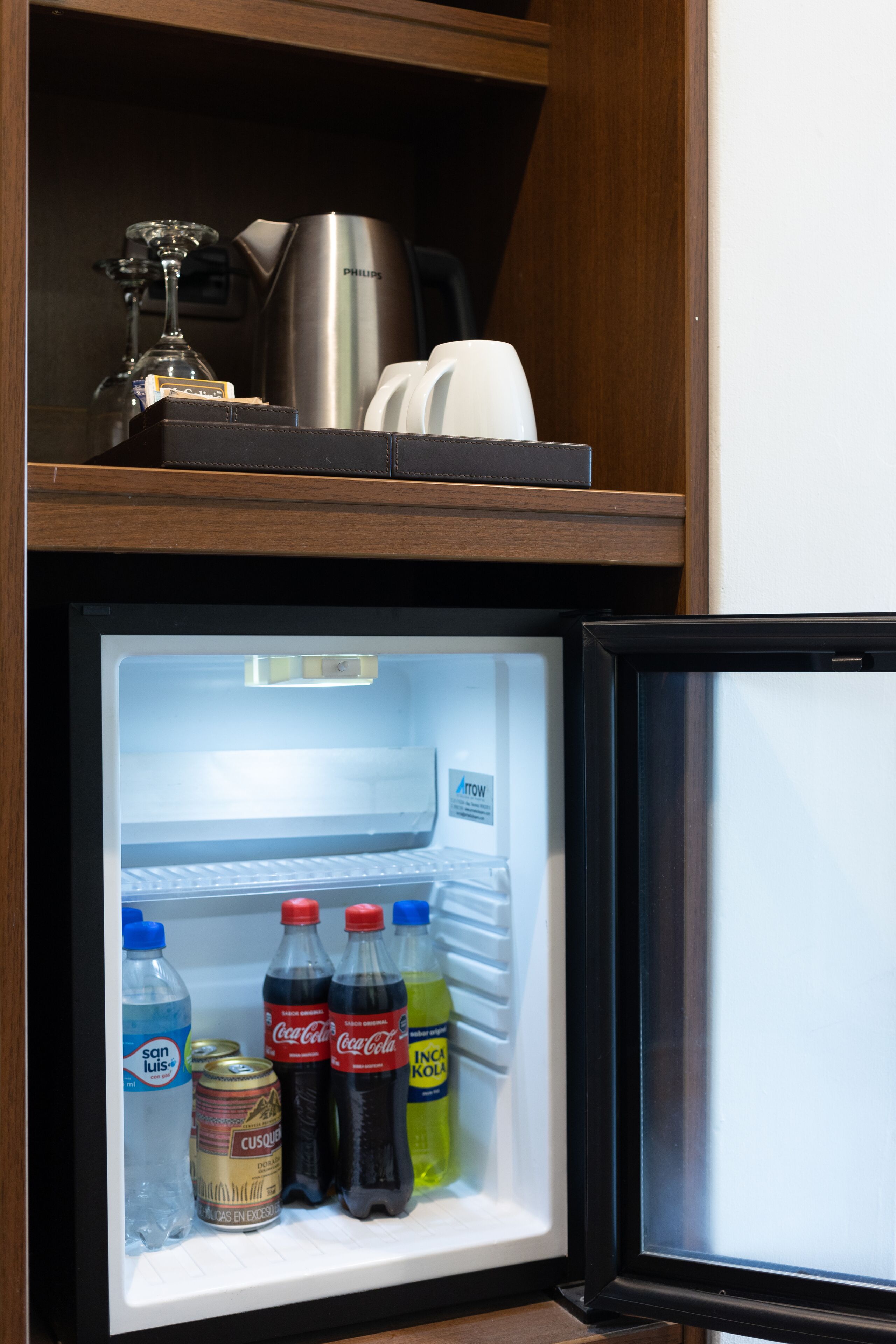 Un réfrigérateur avec une étagère supérieure où se trouvent une machine à café et des verres. À l’intérieur, on trouve des bouteilles d’eau, des sodas et une boîte de conserve.