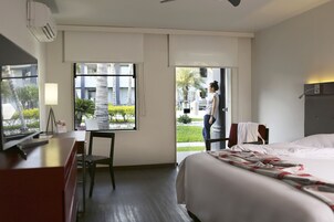 Una habitación de hotel con una cama, un escritorio con una lámpara, una silla y una ventana con vistas a un jardín.