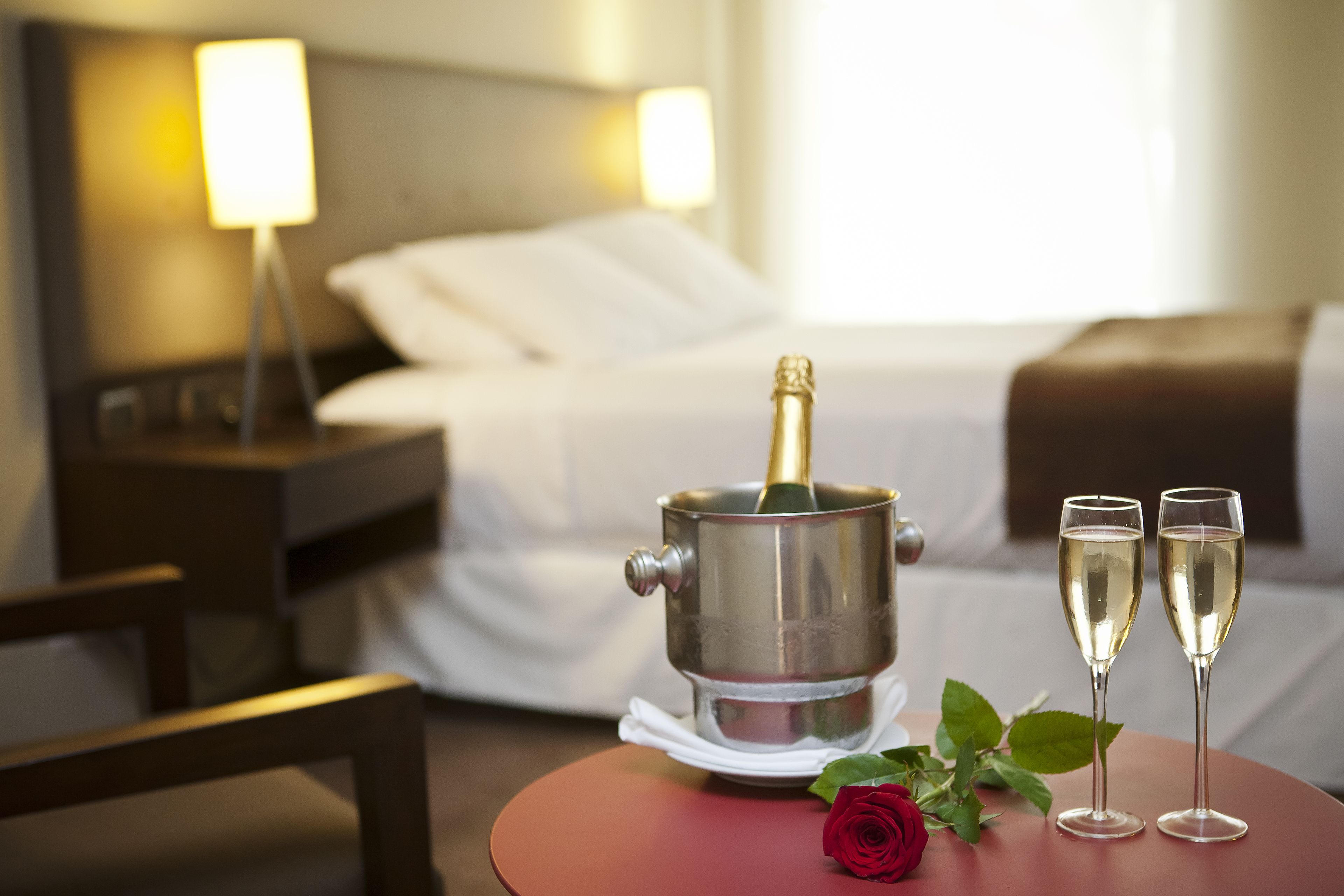 A hotel room with a bed, bedside table, and a table with a champagne bottle and two glasses.
