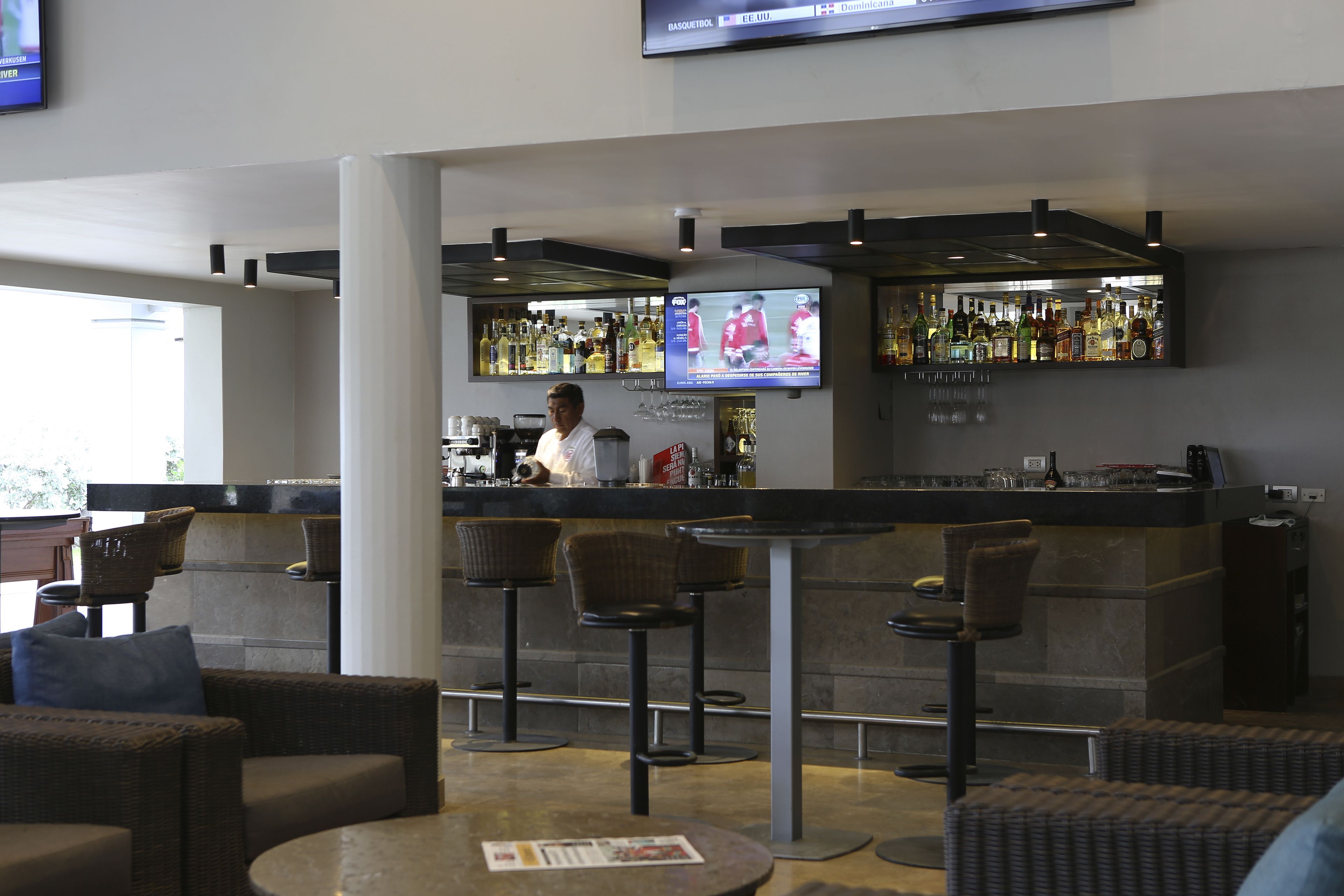 A bar area with wicker furniture, a television mounted above the bar, and a variety of bottles on shelves.