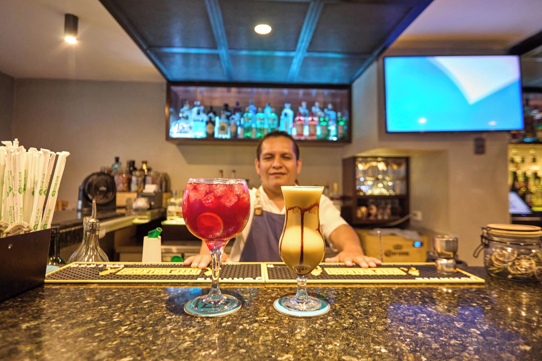 A bar area with two cocktails on the counter, a bartender, and a television screen in the background.