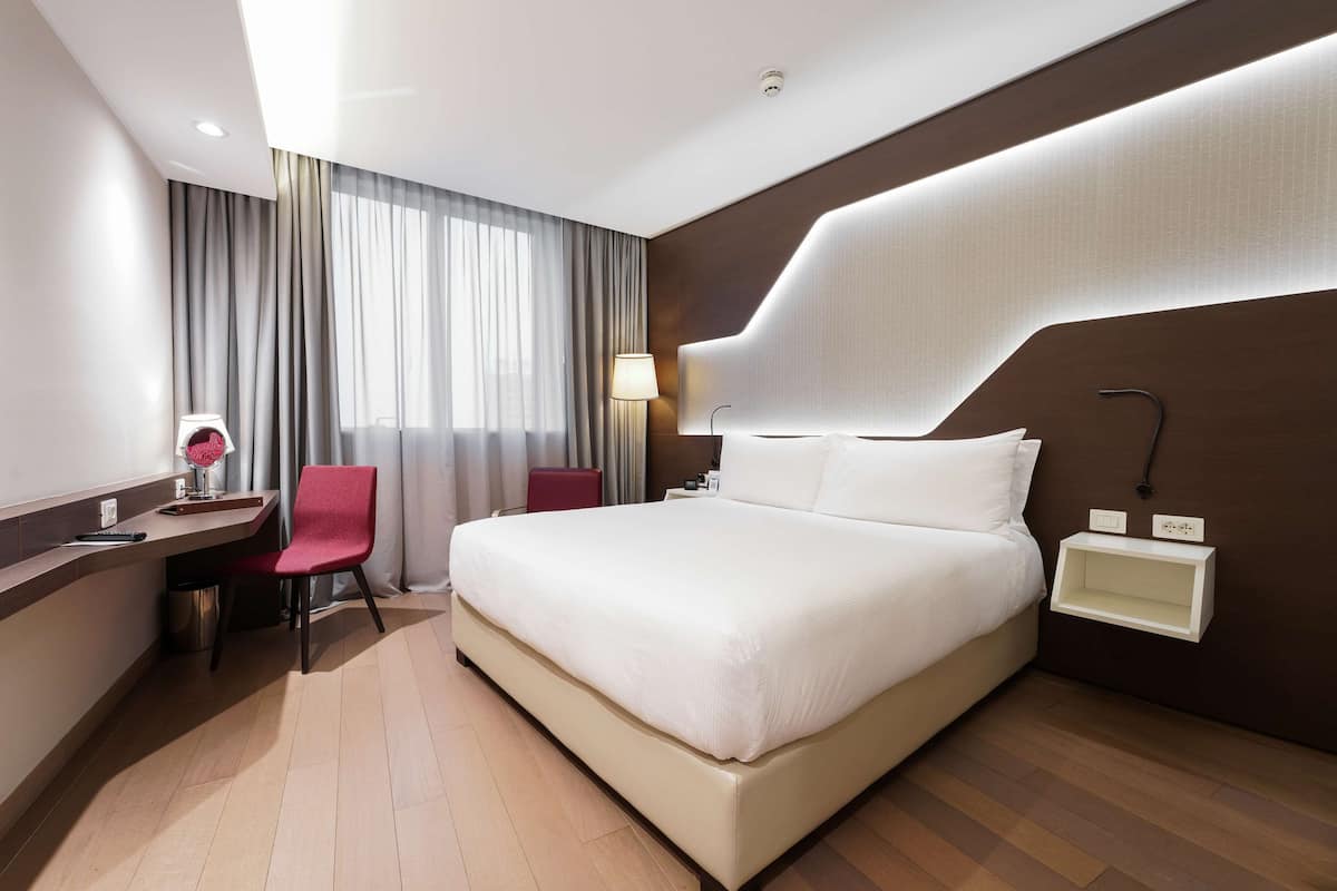 Ein modernes Hotelzimmer mit einem großen Bett, einem Schreibtisch mit Stuhl, einer Lampe und einem Nachttisch.