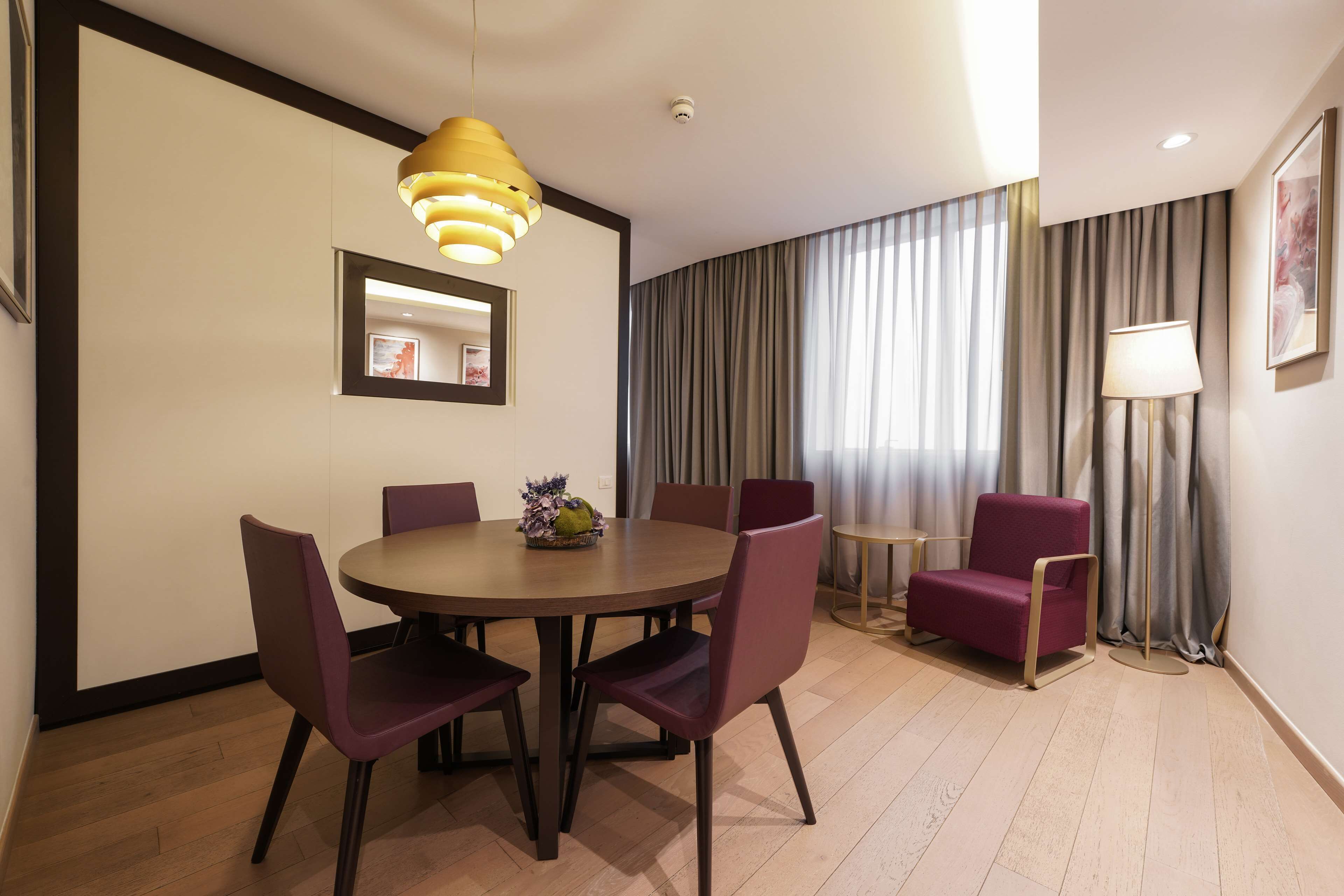 A modern dining area with a round table, purple chairs, a yellow pendant light, and a view of a curtain-covered window.