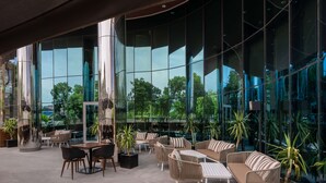 Un espace extérieur moderne, aménagé avec des meubles en osier et des plantes en pot, de grandes baies vitrées réfléchissantes et une vue sur la verdure.