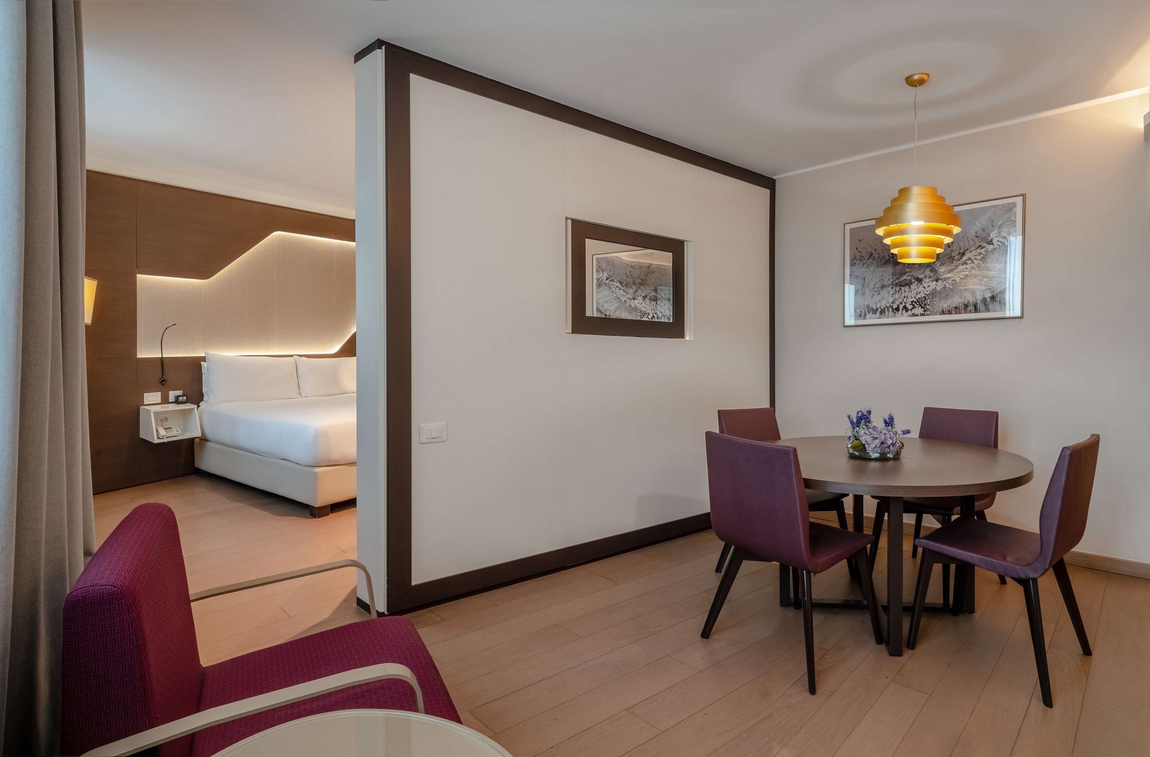 A modern hotel room with a bed, bedside table, and a dining area with chairs and a table.